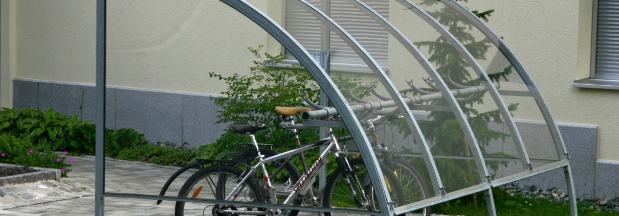Zwei Fahrräder unter einer modernen Fahrradüberdachung aus Metall und transparentem Kunststoff vor einem Wohngebäude